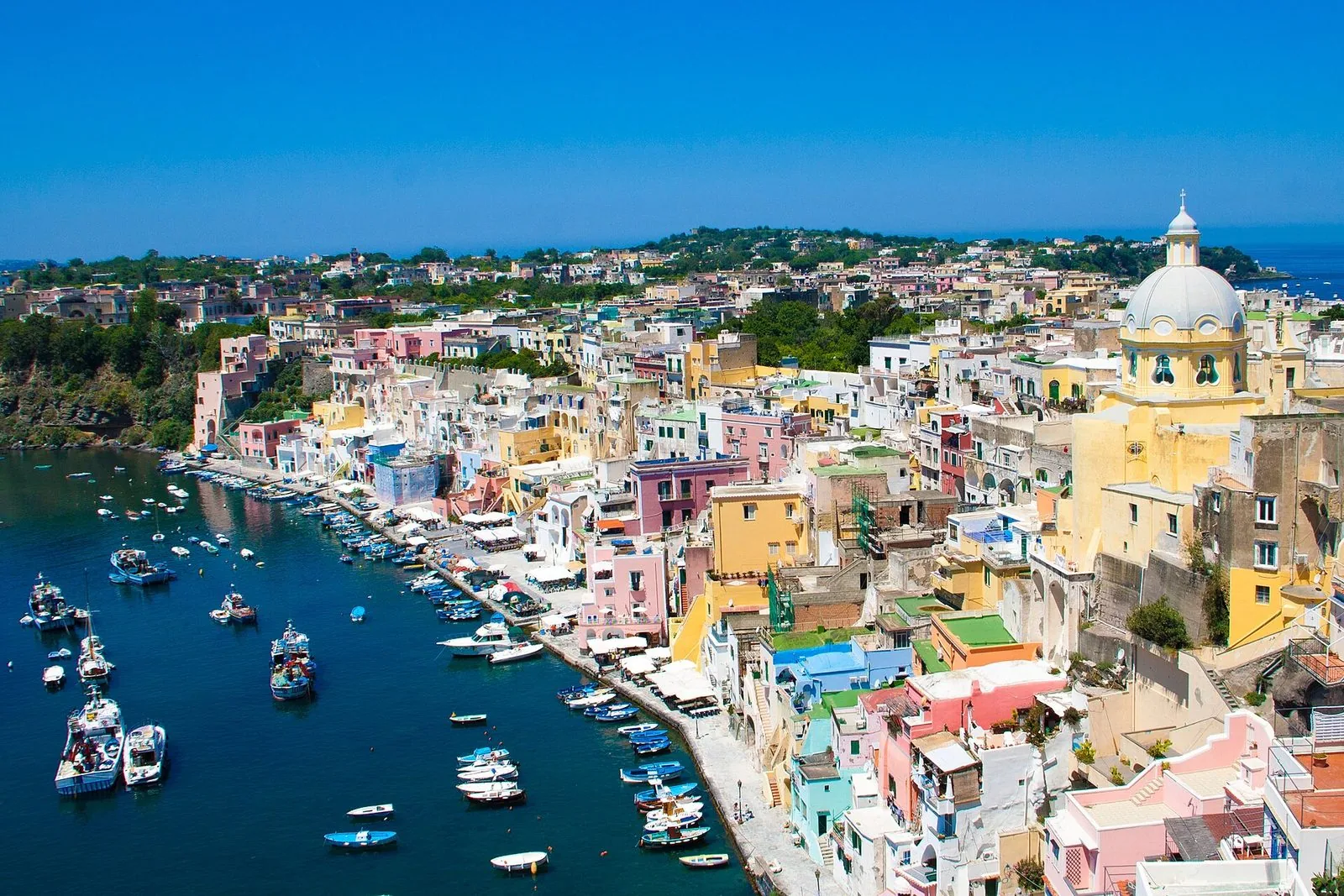 Veduta dall'alto della Marina Corricella a Procida: case color pastello affacciate sul porticciolo, barche ormeggiate nell'acqua blu, una chiesa con cupola chiara a destra e il mare aperto sullo sfondo
