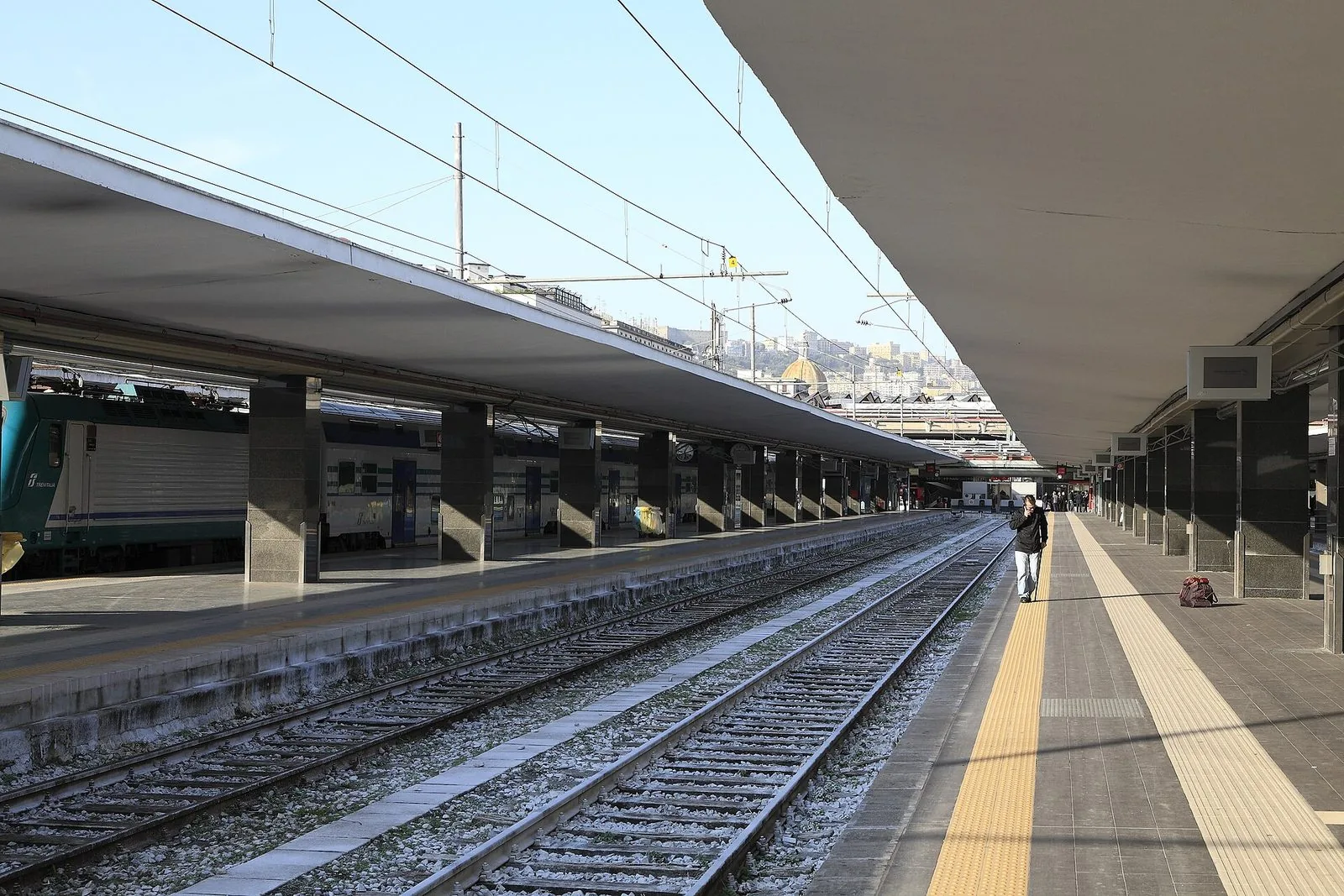 I binari e le lunghe pensiline della stazione di Napoli Centrale in una giornata limpida, con un treno regionale fermo a sinistra e sullo sfondo le case sulla collina e una cupola