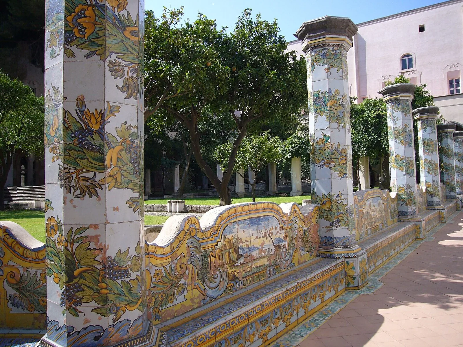 Il chiostro maiolicato di Santa Chiara a Napoli: pilastri ottagonali e sedute rivestiti di maioliche gialle e azzurre con tralci e scene di paesaggio, alberi di agrumi nel giardino e un edificio rosa sullo sfondo