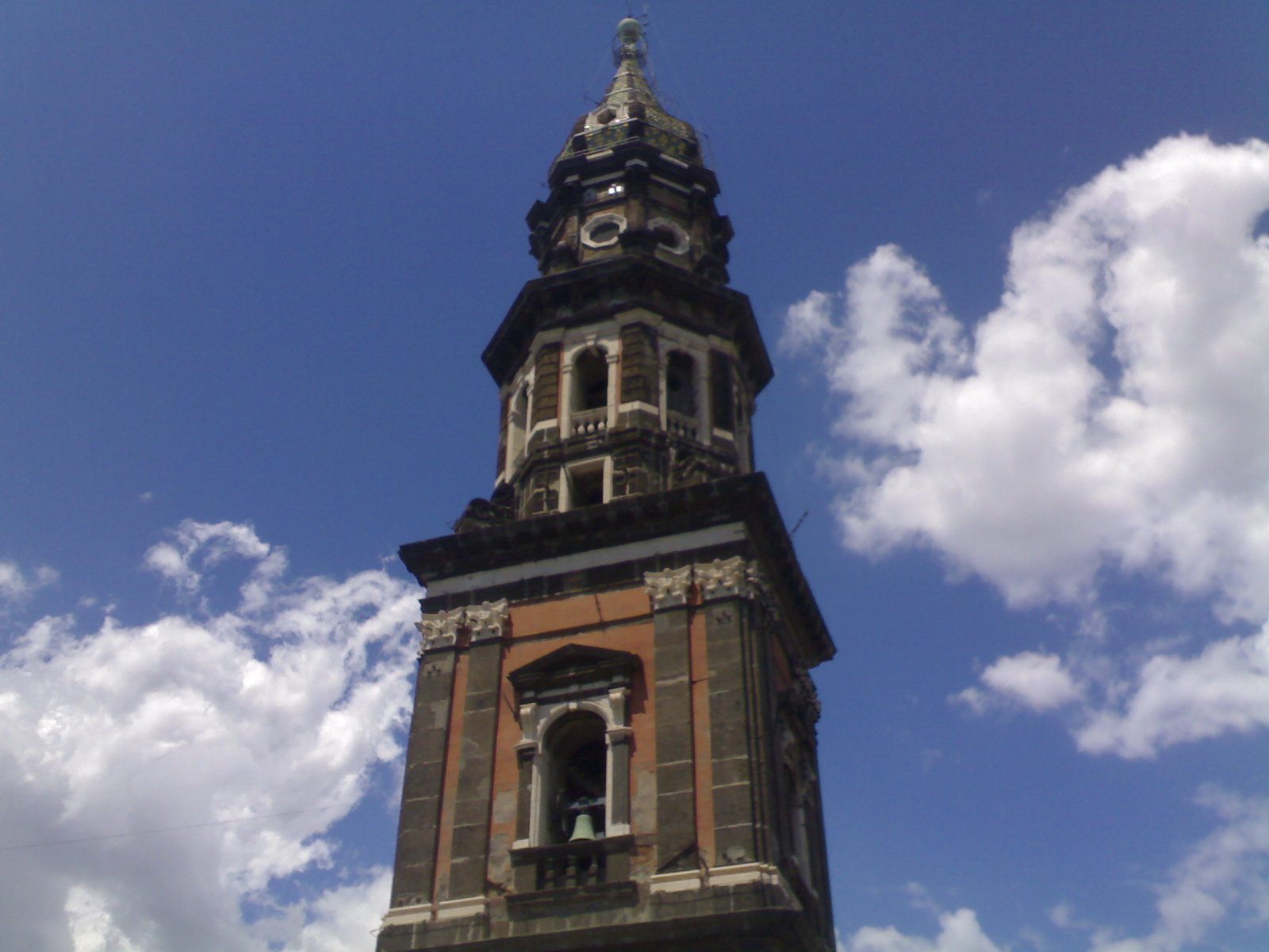 Il campanile della Basilica del Carmine Maggiore a Napoli visto dal basso: torre in pietra scura e mattoni con cella campanaria, ordini sovrapposti e cuspide rivestita di maioliche, contro un cielo azzurro con nuvole bianche