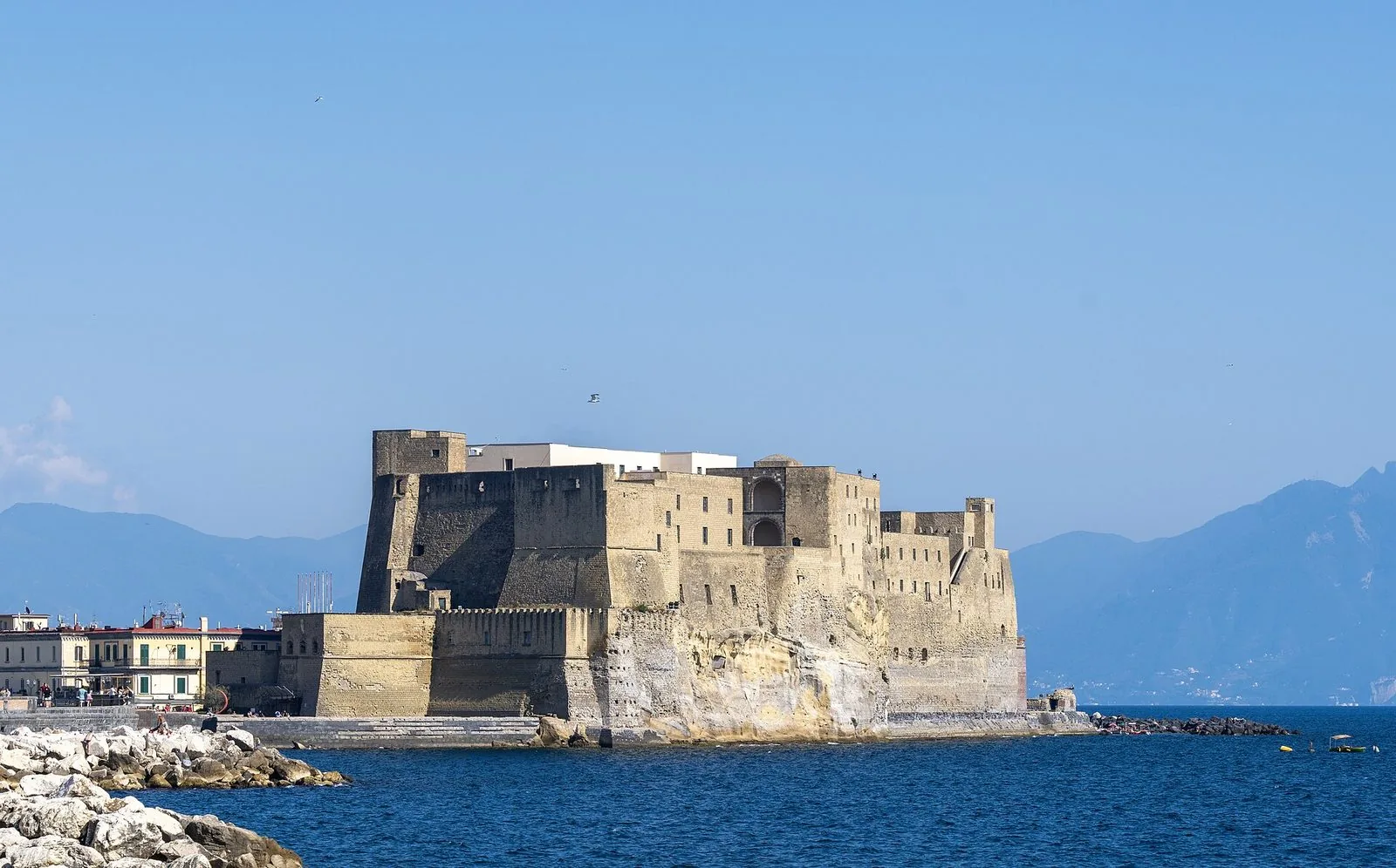 Il Castel dell'Ovo visto dal mare in una giornata limpida: la mole di tufo color sabbia sull'isolotto, le mura merlate, il golfo blu in primo piano e le montagne della penisola sorrentina sullo sfondo