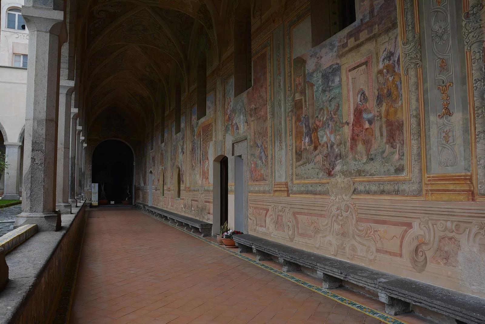 Porticato del chiostro di Santa Chiara a Napoli: pavimento in cotto, panche in pietra lungo la parete e pareti interamente affrescate con scene e cornici decorative, colonne a sinistra verso il giardino