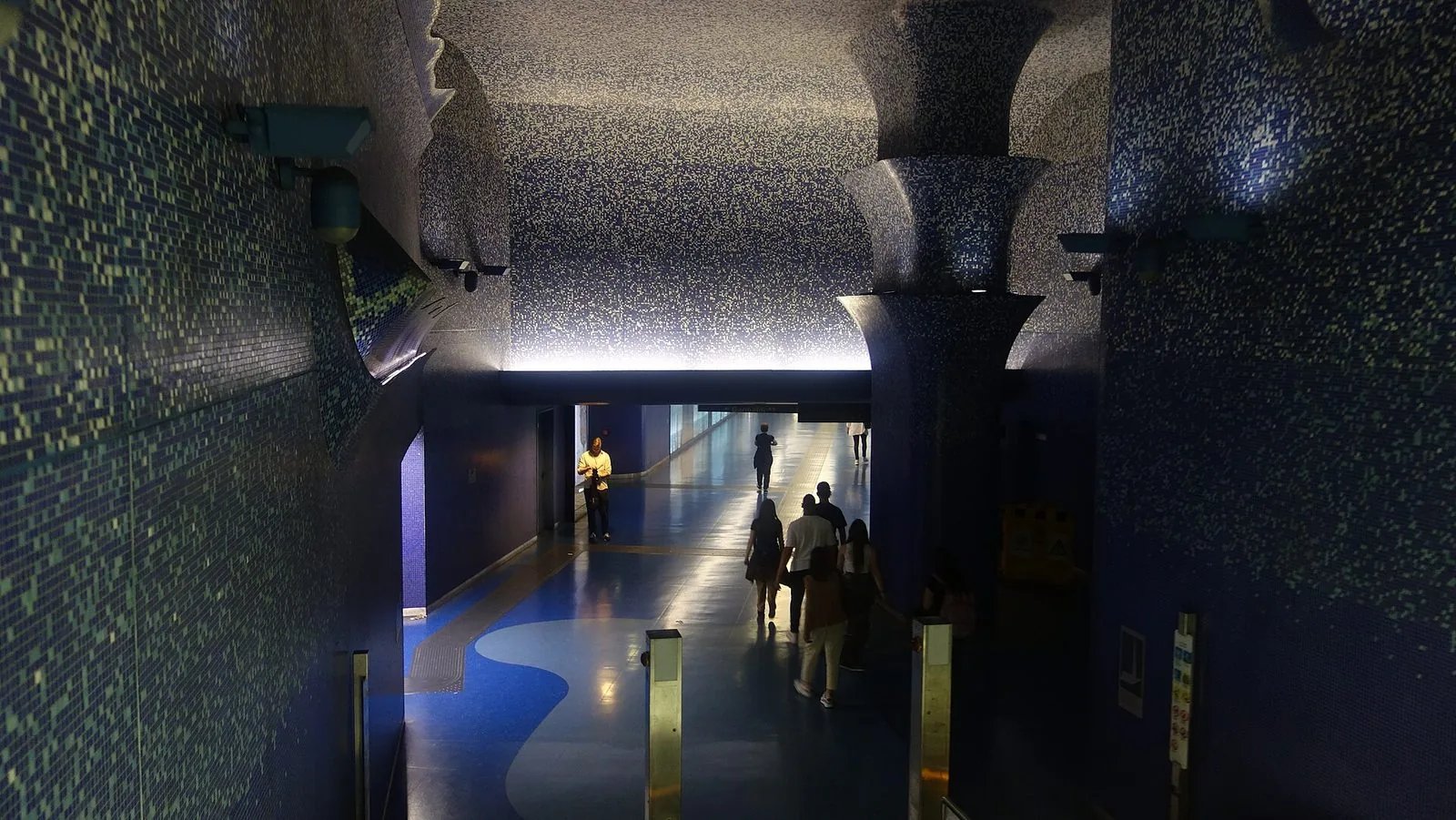 Corridoio della stazione Toledo della metropolitana di Napoli rivestito di mosaici blu e azzurri, con il soffitto luminoso, un grande pilastro decorato e alcuni passeggeri di spalle in lontananza