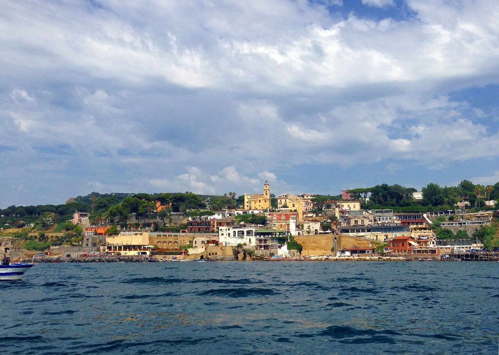 Il borgo di Marechiaro visto dal mare: case colorate e terrazze sul costone tufaceo di Posillipo, un campanile tra i pini e la piccola spiaggia affollata, sotto un cielo di nuvole bianche