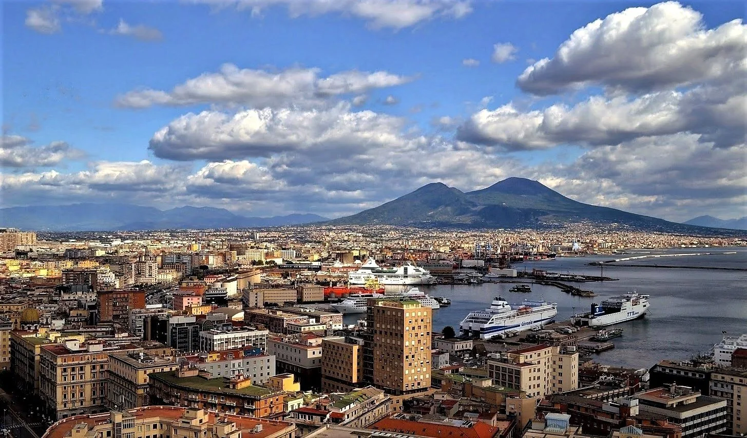 Panorama di Napoli dall'alto sotto un cielo azzurro con grandi nuvole bianche: i palazzi della città, il porto con traghetti e navi da crociera e, sullo sfondo, il Vesuvio